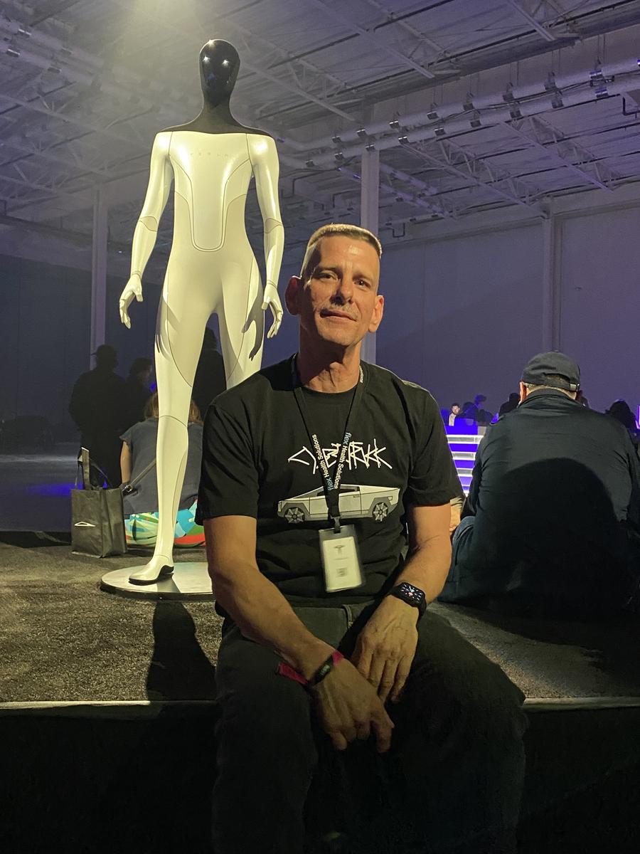 Gary D Murphy seated in front of Tesla Optimus humanoid robot under moody blue stage lighting